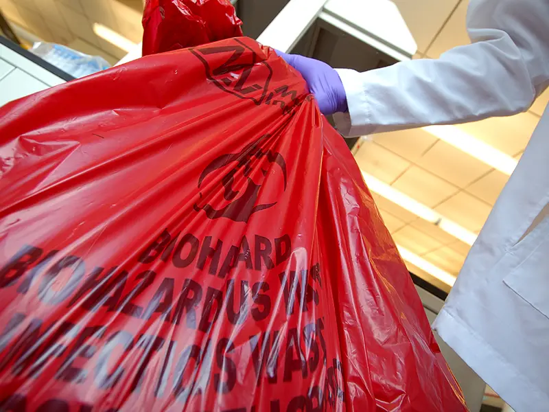 Person in lab coat holding a red biohazard medical waste bag.
