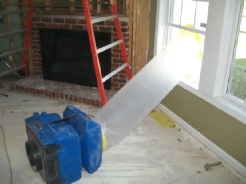 Blue air scrubber with a clear plastic duct attached, positioned near a window in a room under renovation.