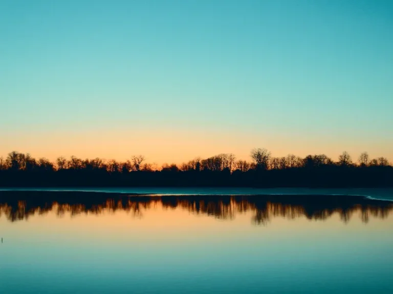 Calm lake reflecting a tree-lined shore at sunset with a clear sky.