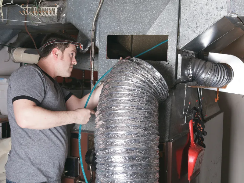 A man wearing a headlamp inspects a large flexible duct connected to a metal HVAC unit.