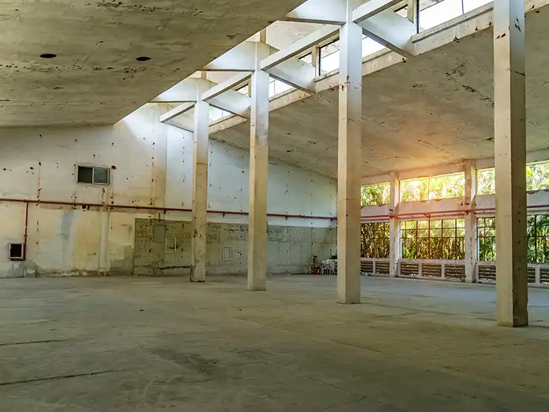 Empty industrial warehouse interior with concrete pillars and sunlight streaming through windows.