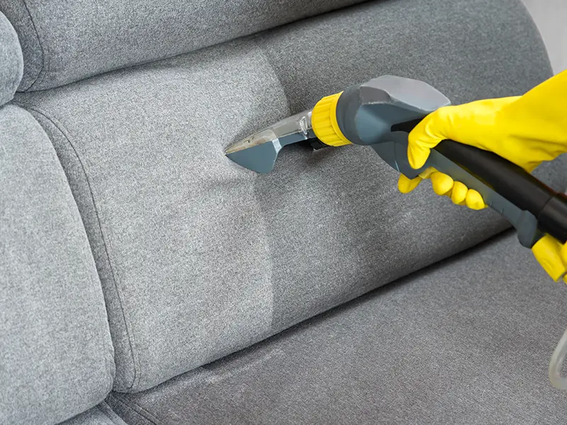 Hand in yellow glove using a steam cleaner on a gray fabric sofa.