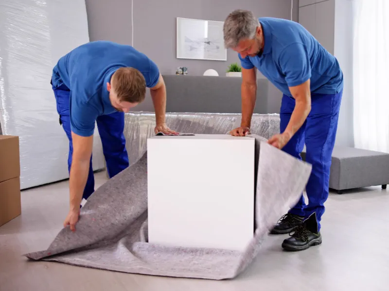 Two men in blue shirts wrapping a white box with protective fabric indoors.