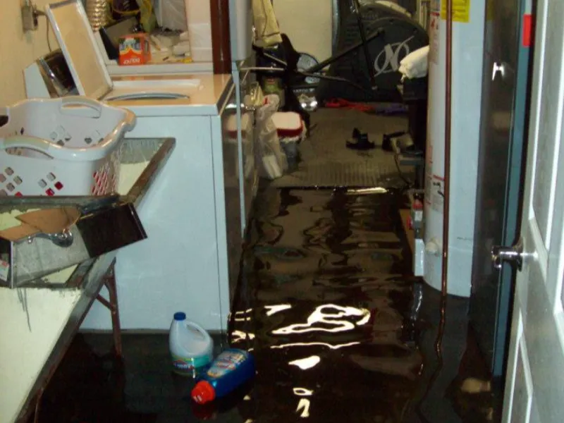 Water flooding the floor of a laundry room with a washing machine and cleaning supplies.